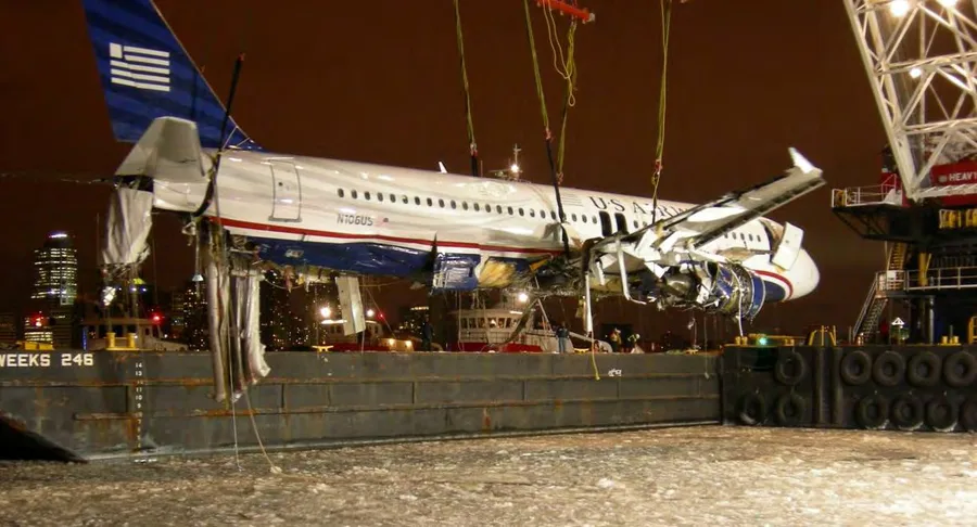 The wreckage of N106US being lifted by crane onto a barge at night, with the Manhattan skyline in the background